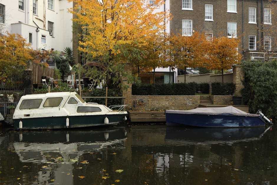 A tranquil riverside scene featuring two boats moored along a canalbank, with one boat displaying a weathered white and green hull and the other a sleek dark blue with a protective cover. Behind the b